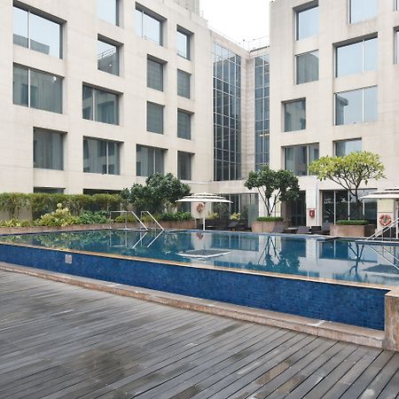 Holiday Inn New Delhi Aerocity By Ihg Exterior foto The photo shows a modern outdoor swimming pool situated in a courtyard surrounded by several buildings. The pool has a sleek design with a blue tiled edge and is bordered by wooden decking. There are a few trees and plants around the area, adding som