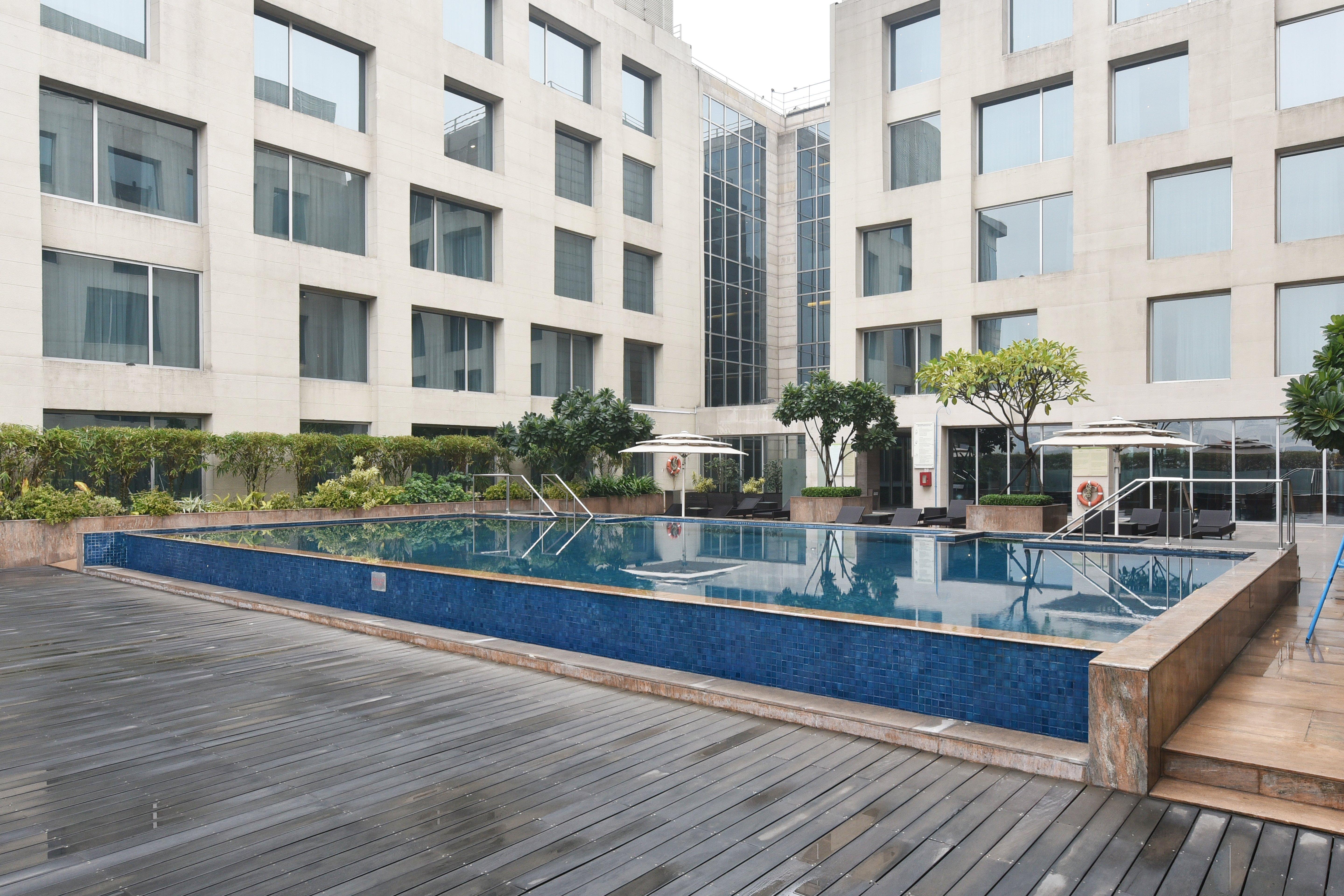 Holiday Inn New Delhi Aerocity By Ihg Exterior foto The photo shows a modern outdoor swimming pool situated in a courtyard surrounded by several buildings. The pool has a sleek design with a blue tiled edge and is bordered by wooden decking. There are a few trees and plants around the area, adding som
