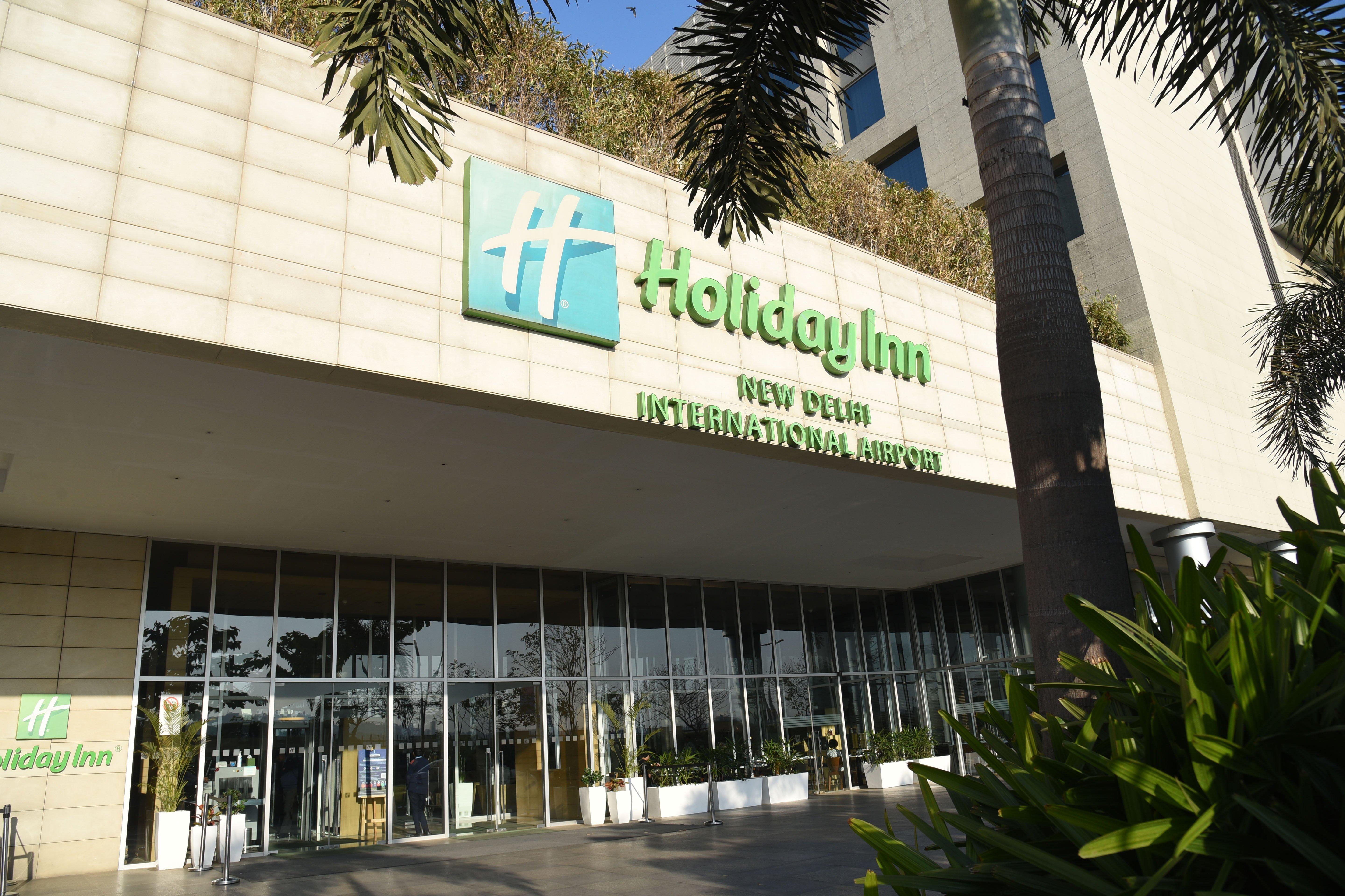 Holiday Inn New Delhi Aerocity By Ihg Exterior foto The photo shows the exterior of a Holiday Inn hotel, specifically the Holiday Inn New Delhi International Airport. The building features a modern design with a prominent green and white sign displaying the hotel name. There are large glass doors at t