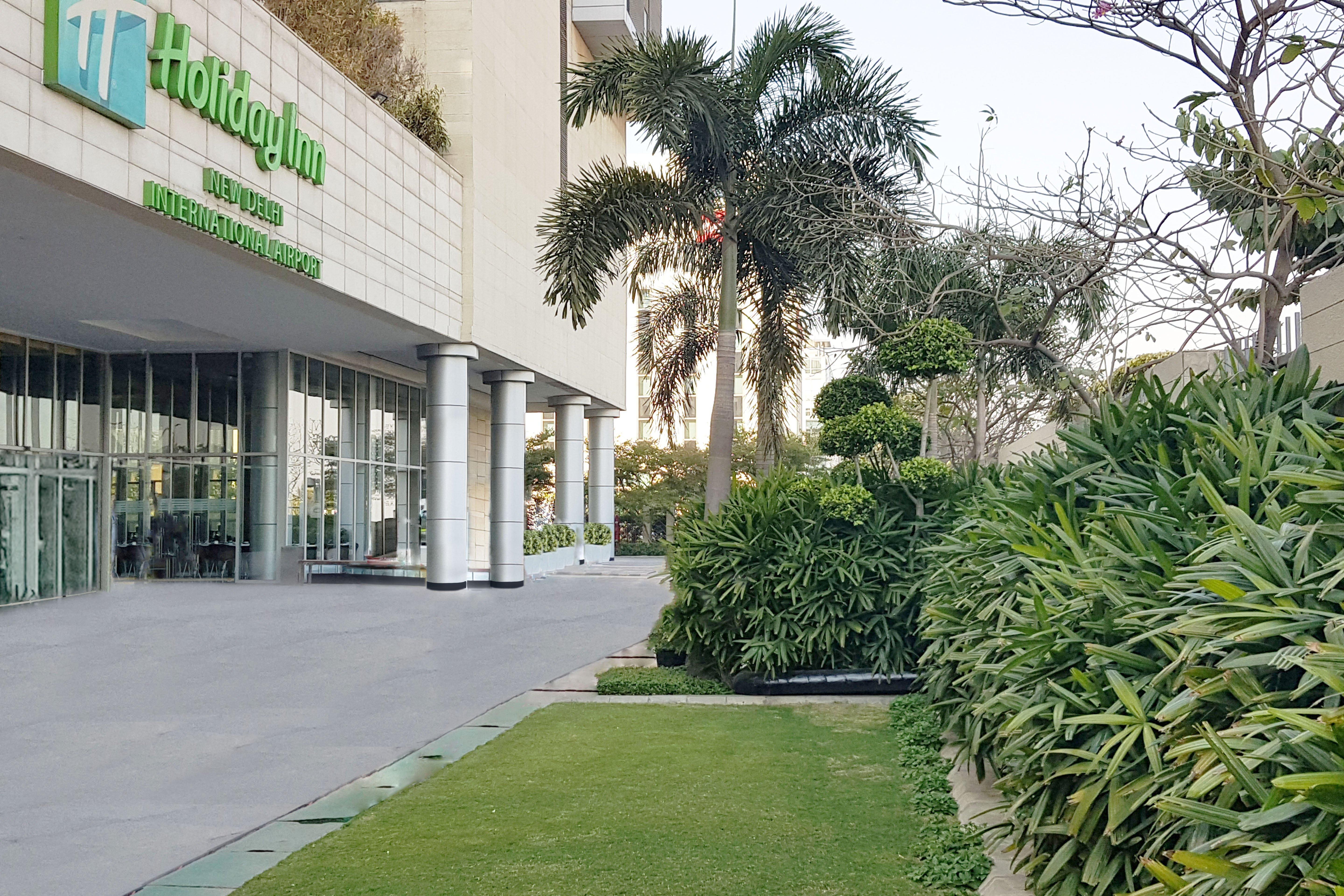 Holiday Inn New Delhi Aerocity By Ihg Exterior foto The photo shows the exterior of a Holiday Inn hotel. In the foreground, there is a well-maintained green lawn and various ornamental plants, creating a pleasant garden area. The building features large glass doors and columns, and the hotel's name an